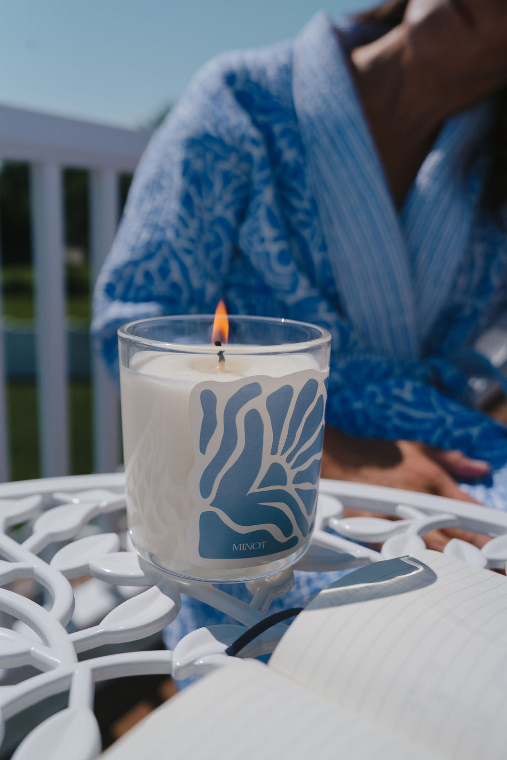 Lit candle in a glass holder with a blue abstract design, placed on a white metal table. A person in a blue robe is sitting in the background.