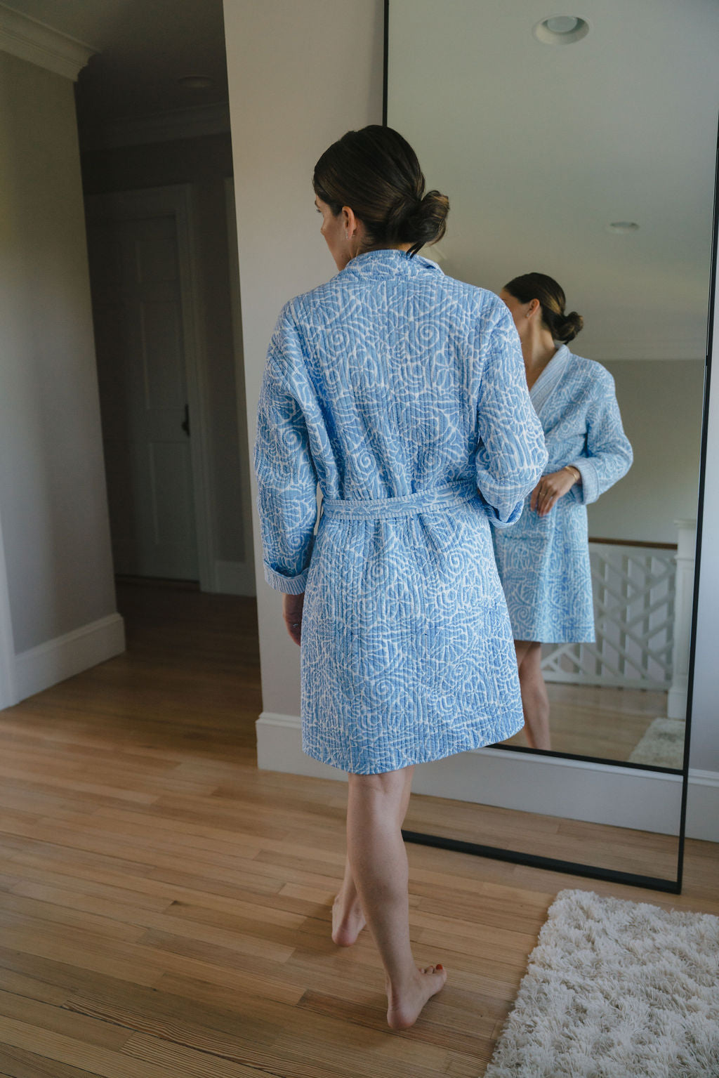 Woman in a baby blue patterned cotton robe standing barefoot in front of a mirror on a wooden floor.