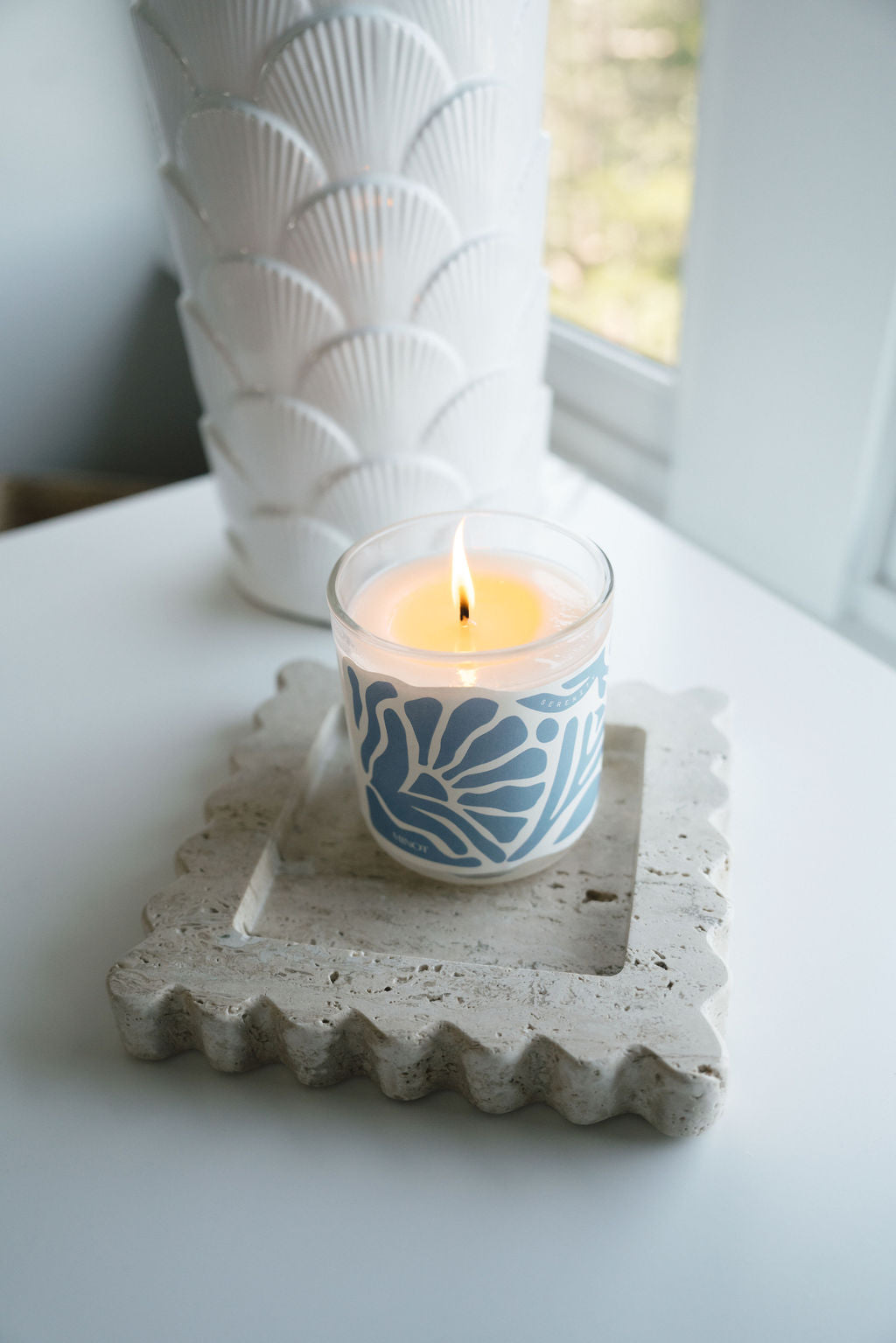 A lit candle with a blue and white coastal design sits on a textured, square stone coaster. Behind it is a white vase with a scallop pattern, near a window with soft daylight.