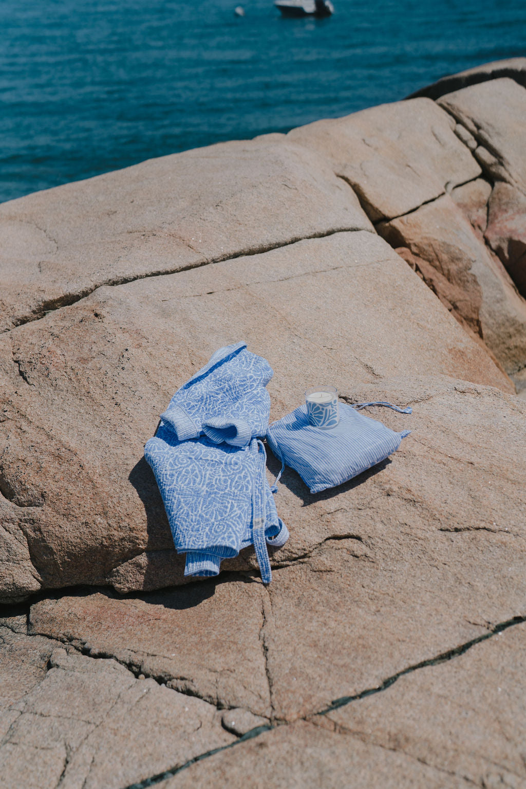 Baby blue patterned cotton robe, matching bag, and candle lie on sunlit rocks by the sea, with a boat visible in the background.