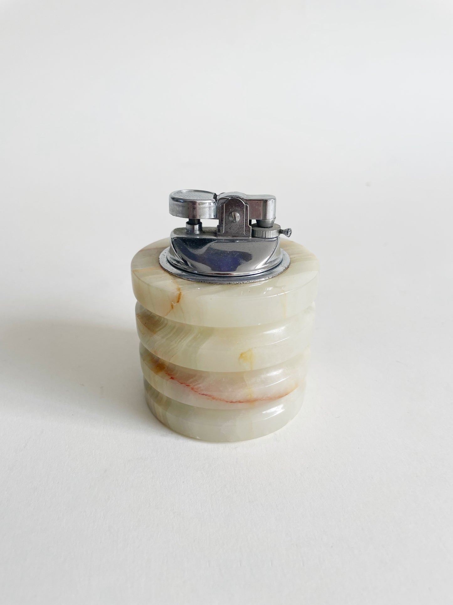 A round, ridged stone lighter with a metal top, placed on a white surface. The stone has natural beige tones with subtle streaks of color.