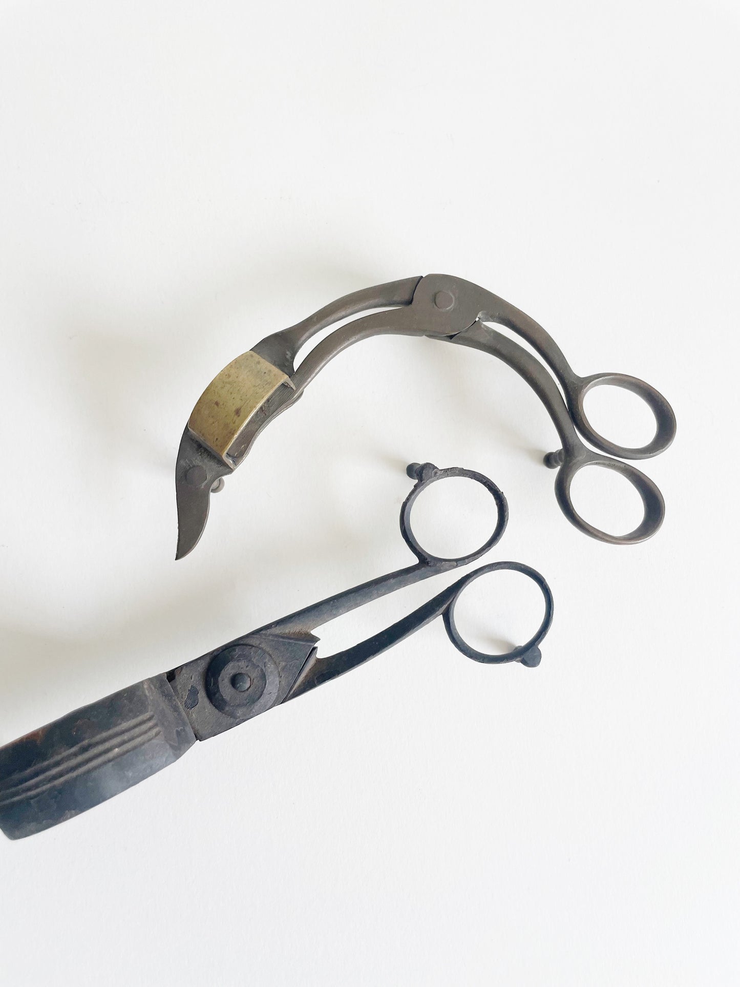 Two vintage wick trimmers on a white background. One is curved with a brass detail, and the other is straight with circular finger holes. Both have an aged, metallic finish.