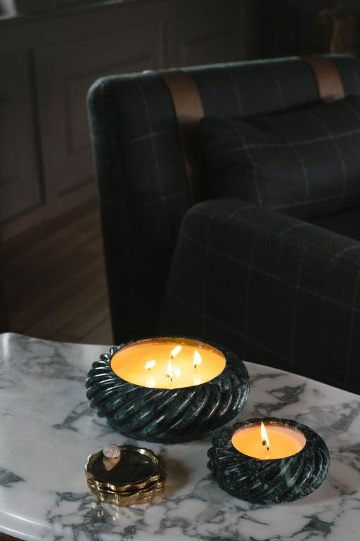 Two lit candles in ornate emerald stone holders on a marble table, with a small decorative object nearby. The background features a dark, plaid-upholstered couch.