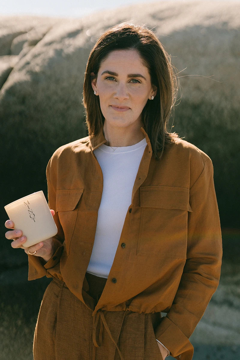 MINOT's founder Kerry Kip stands outdoors holding a MINOT candle. She's wearing a brown outfit and a white top, with large rocks in the background under a clear sky.