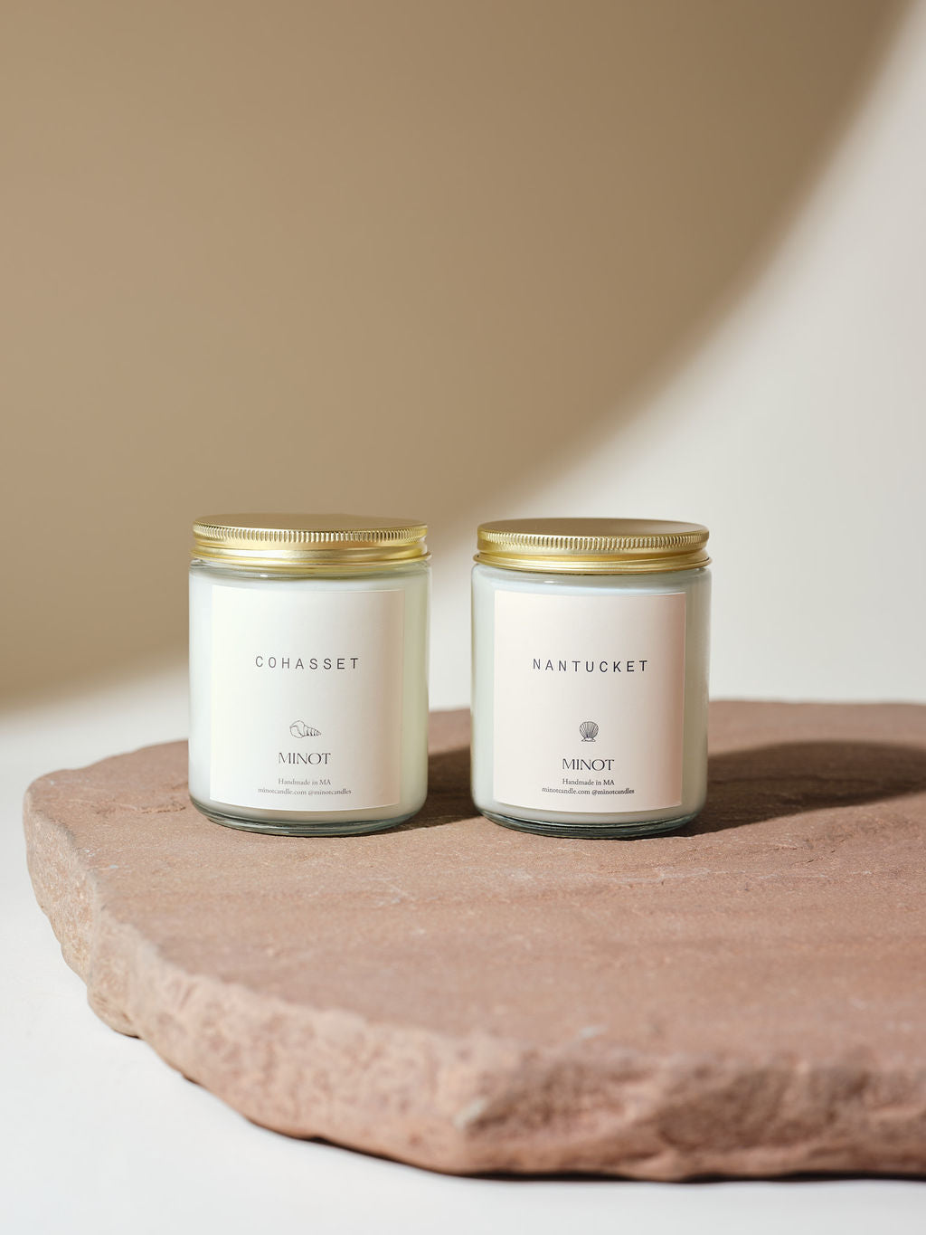 Two soy candles in glass jars labeled "Cohasset" and "Nantucket" with gold lids sit on a flat stone surface against a simple, neutral background.