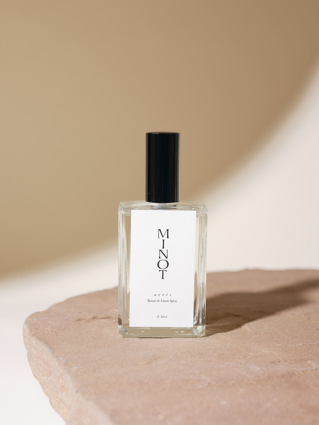 An elegant glass bottle of MINOT Aeris Room & Linen Spray sits on a textured stone surface. The label is white with black text, and the cap is black. The background is softly lit in neutral tones.