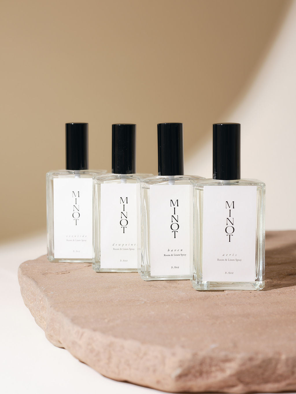 Four clear glass bottles of MINOT Room & Linen Spray with black caps are displayed on a textured stone surface, set against a neutral beige background.