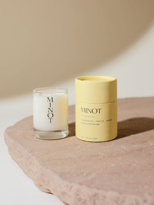 A small soy candle in a glass holder labeled "MINOT" sits on a stone surface next to a cylindrical yellow cardboard box with text describing the Dewpoint scent as a blend of chamomile, spruce, and amber.