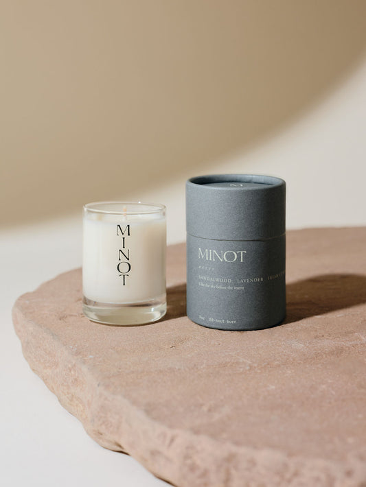 A small white soy candle in a glass holder labeled "MINOT" sits on a stone surface next to a cylindrical blue-gray cardboard box with text describing the Aeris scent as a blend of sandalwood, lavender, and fresh citrus.