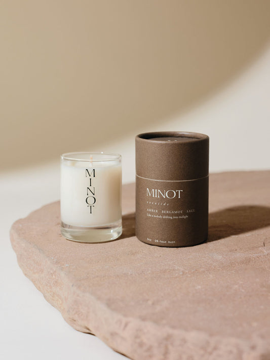 A small white candle in a glass holder labeled "MINOT" sits on a stone surface next to a cylindrical brown cardboard box with text describing the Eventide scent as a blend of amber, bergamot, and sage.