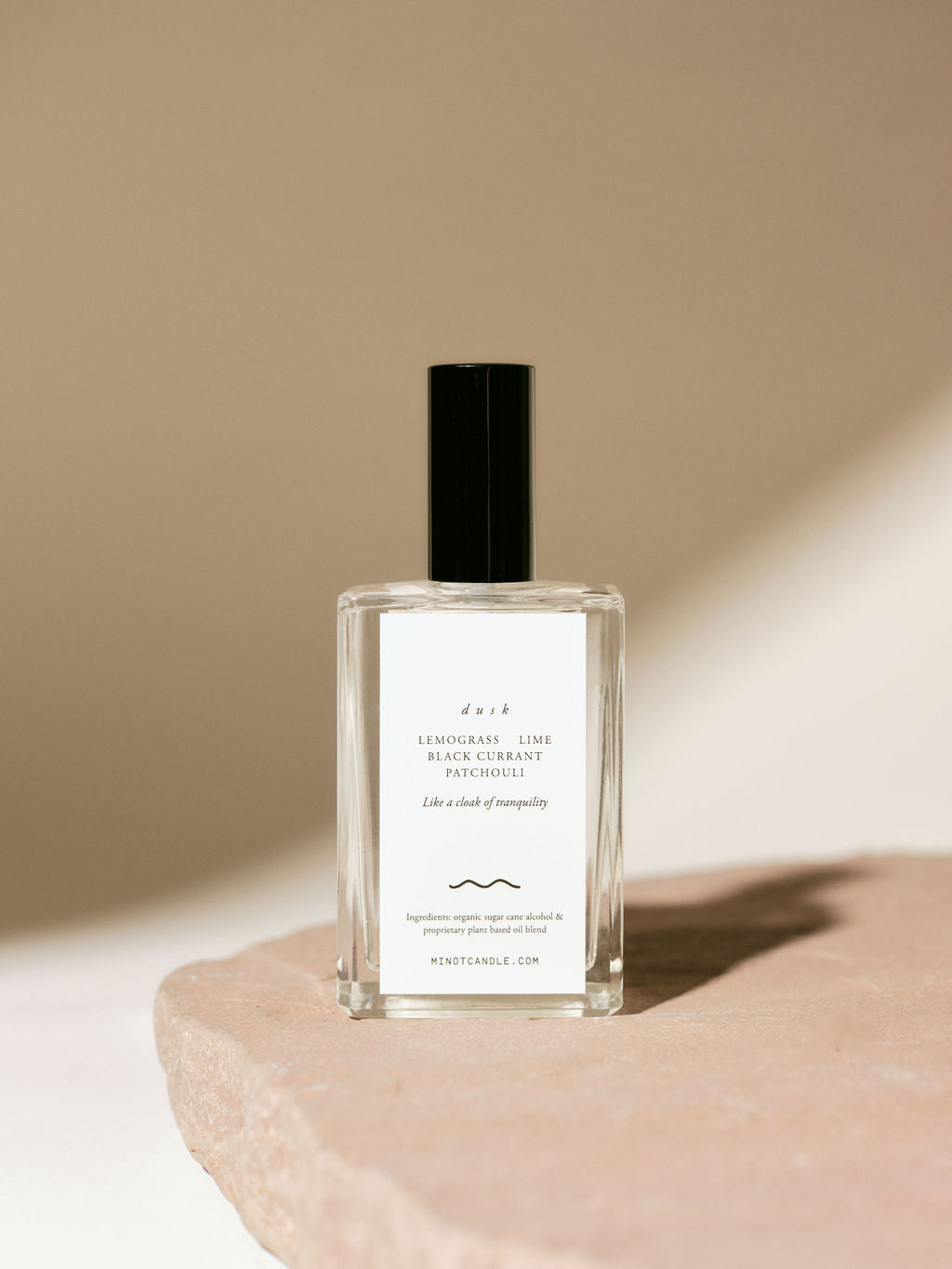 A bottle of MINOT's Dusk room and linen spray with lemongrass, lime, black currant, and patchouli scents sits on a stone surface, with a minimalistic design and soft lighting casting gentle shadows.