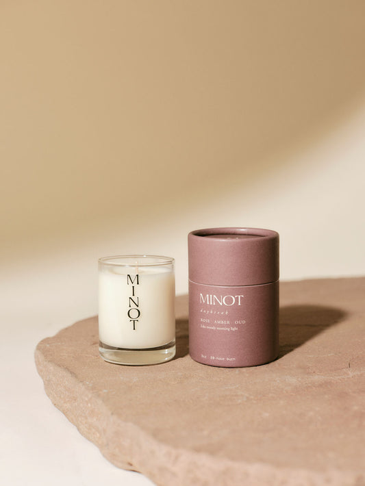 A MINOT Daybreak mini candle in a glass holder is next to a matching cylindrical box on a textured stone surface.