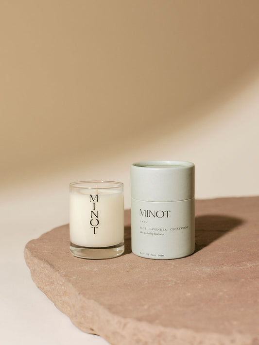 A MINOT Cove mini candle in a glass holder sits beside a light green cylindrical box on a textured stone surface.