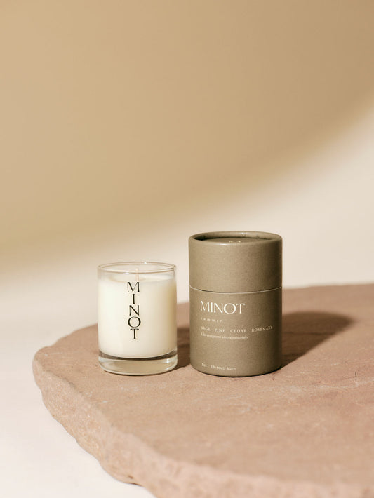 A MINOT Summit mini candle in a glass holder sits beside its cylindrical packaging on a textured stone surface.