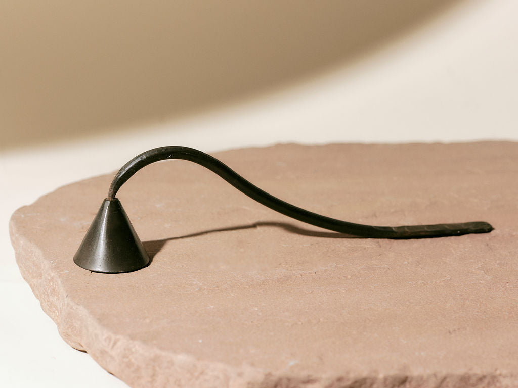 A dark metal candle snuffer with a curved handle resting on a textured, light brown surface.