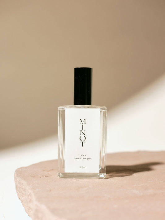 Clear glass bottle of MINOT Cove room and linen spray with black cap, placed on a textured stone surface, highlighted by soft natural light.