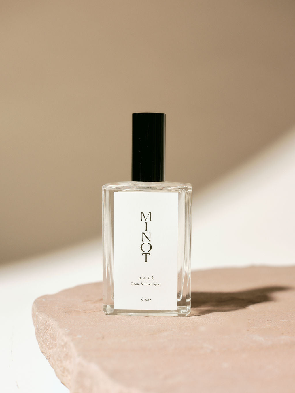 Clear glass bottle of MINOT Dusk room and linen spray with black cap, placed on a textured stone surface, highlighted by soft natural light.