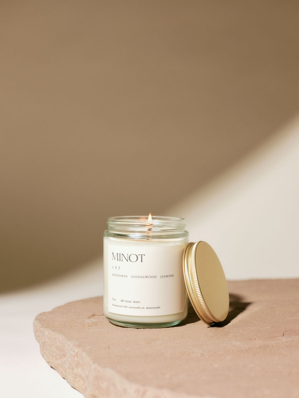 A lit 143 candle in a glass jar labeled "MINOT," placed on a textured stone surface. The jar's label mentions scent notes of mandarin, sandalwood, and jasmine. A golden lid rests beside it.