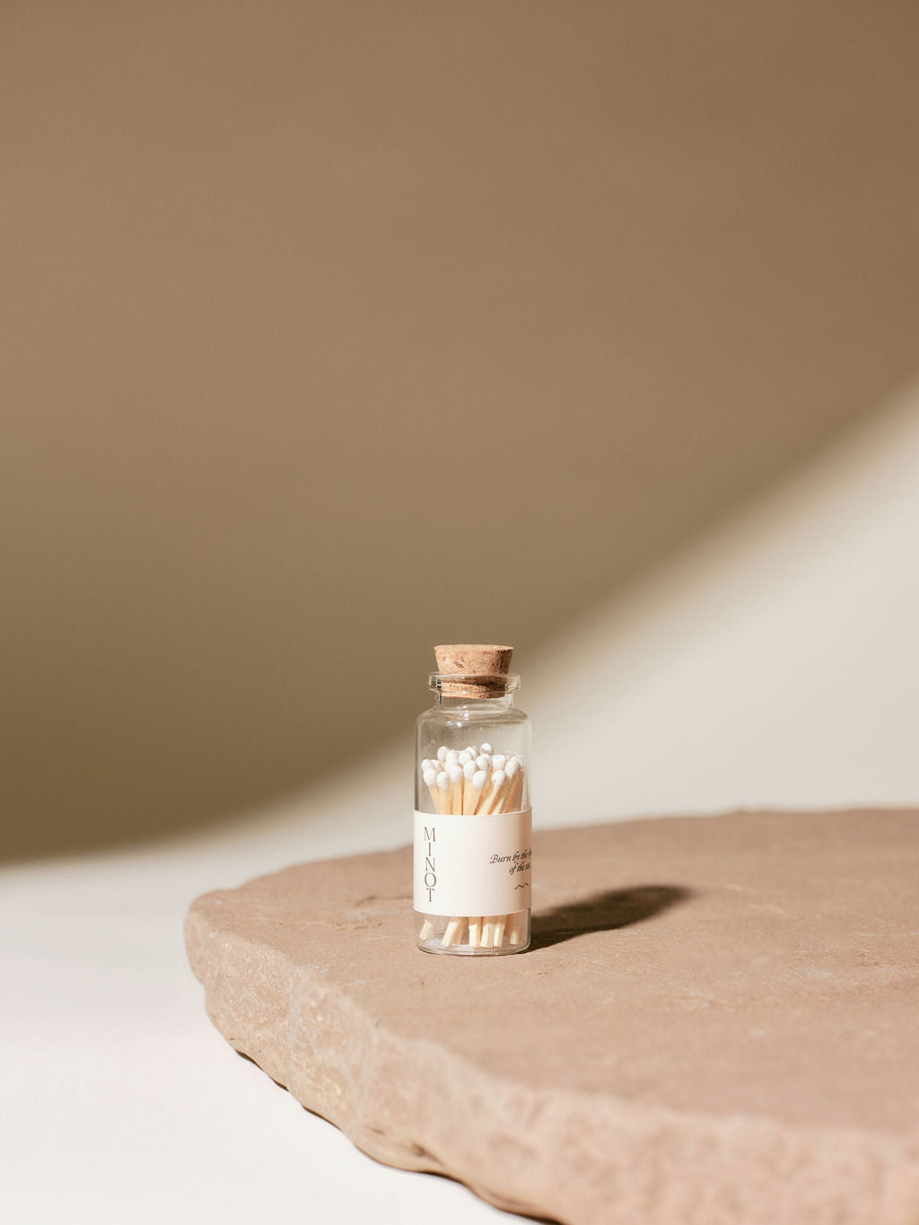 Small glass bottle with a cork lid containing white-tipped matches stands on a textured stone surface, set against a neutral background with soft lighting.