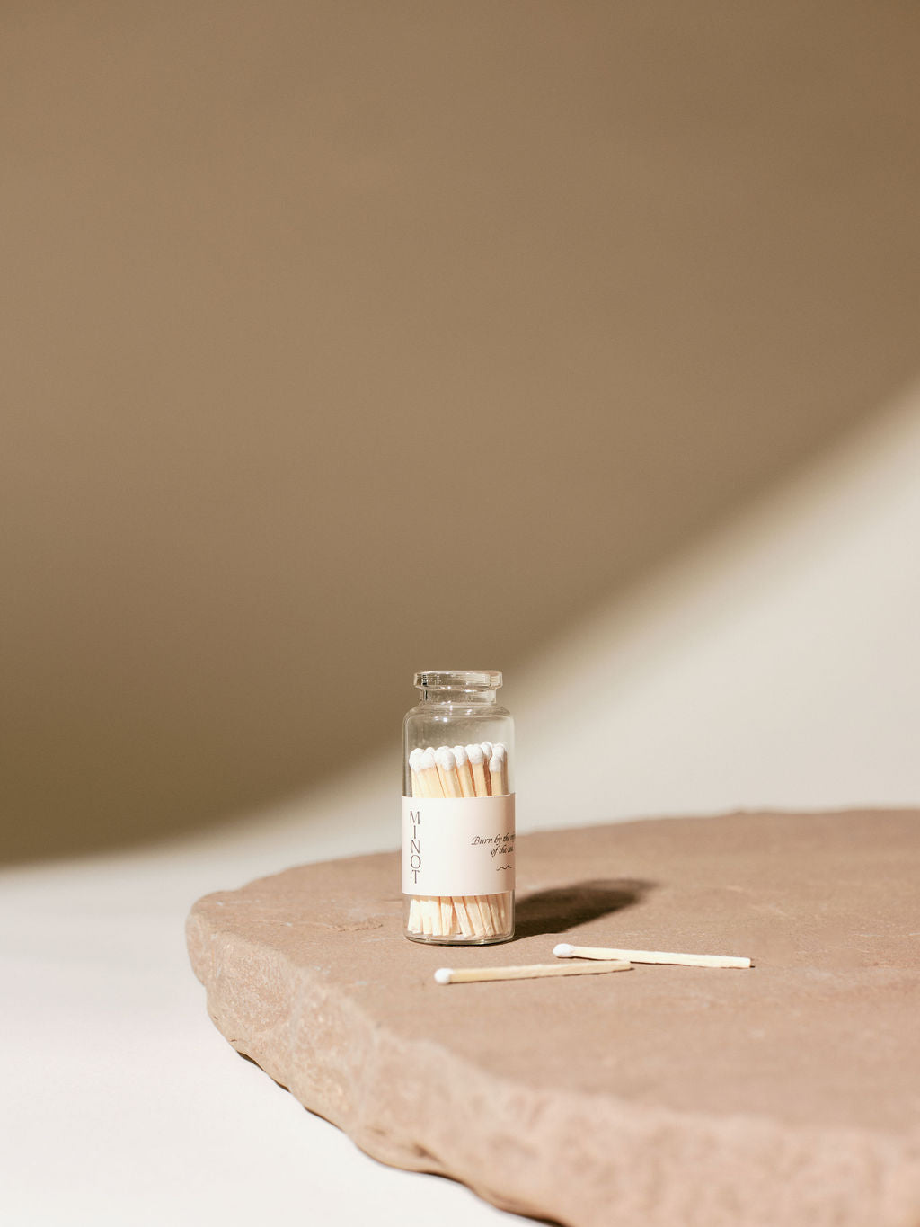 A small glass jar of matches with a white "MINOT" label sits on a textured stone surface. Two matches lie beside the jar. The background is softly lit with shadows.