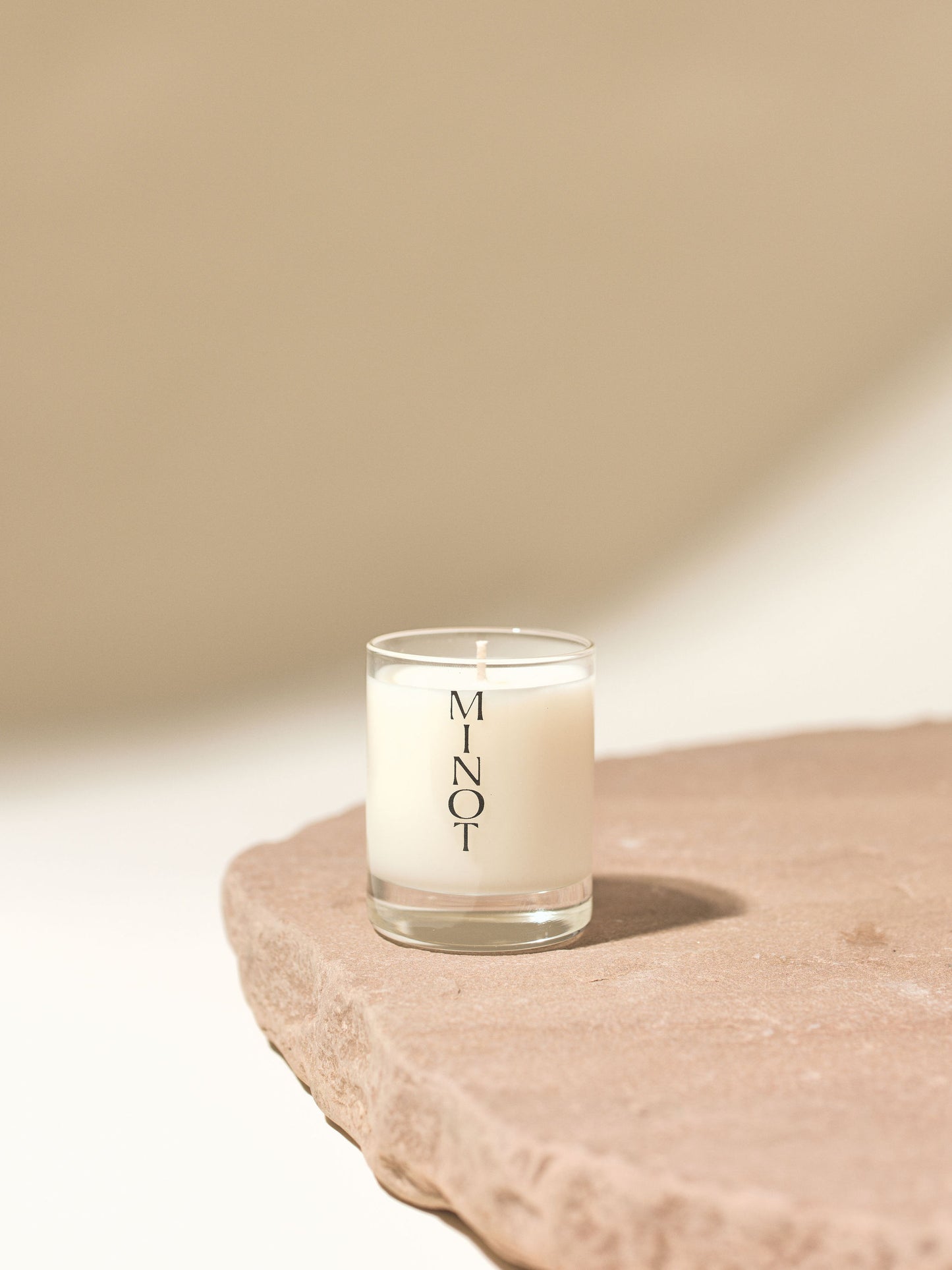 An unlit mini soy candle in a small glass vessel with "MINOT" printed vertically, placed on a textured stone surface against a soft, neutral background.