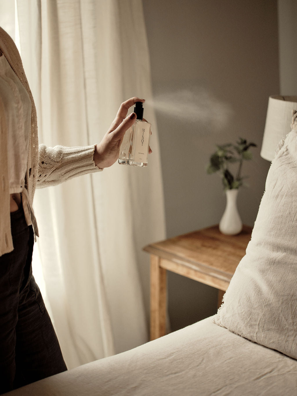 Person spraying MINOT linen and room spray onto a bed with beige linens. A wooden side table with a white vase and green plant is in the background. Light filters through a curtain.