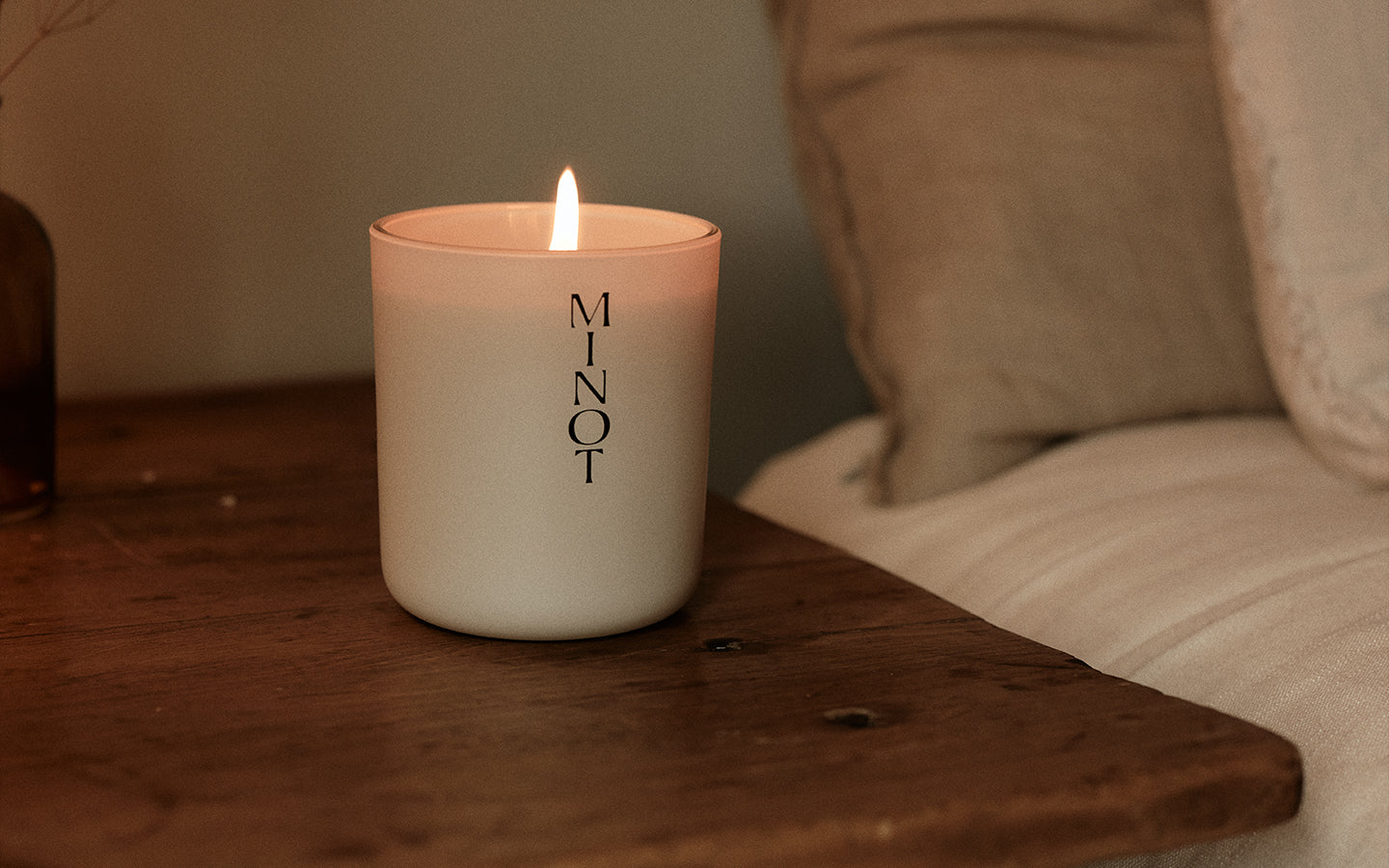 A lit MINOT candle sits on a wooden table next to a beige cushion, creating a cozy atmosphere.