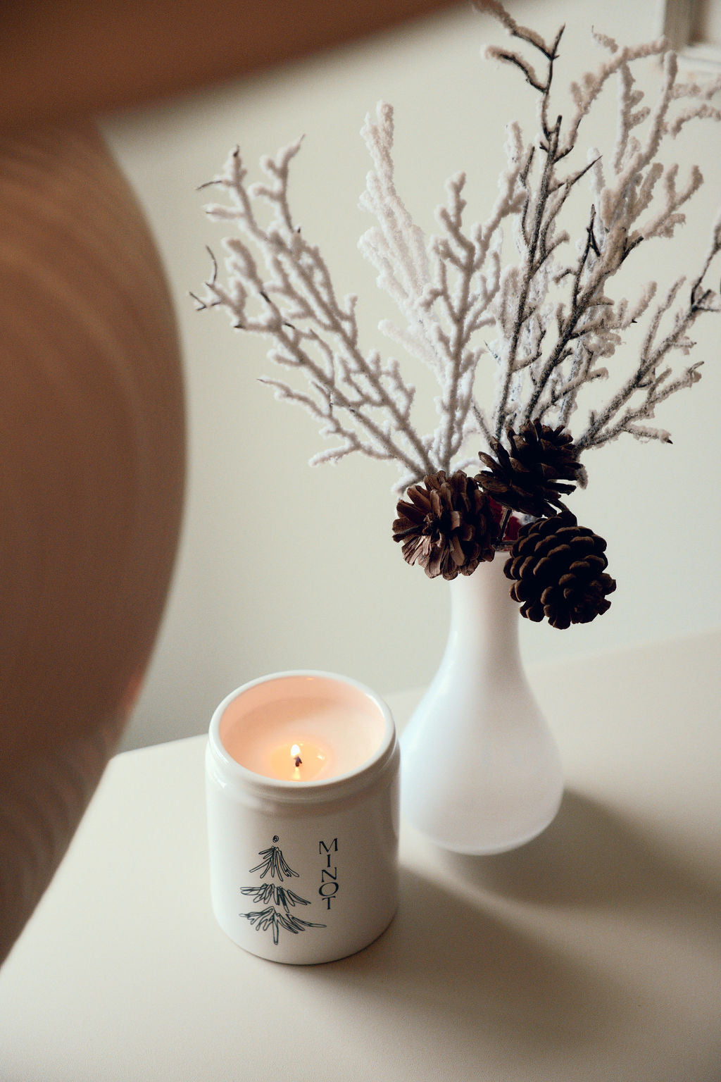 A lit candle in a white jar with "MINOT" and a tree design sits next to a white vase holding pinecones and frosted branches on a light surface.