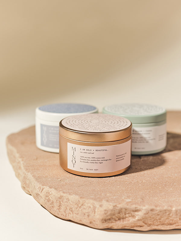 Three affirmation candles in tins with affirmation labels on them placed on a flat stone surface against a soft, neutral background.