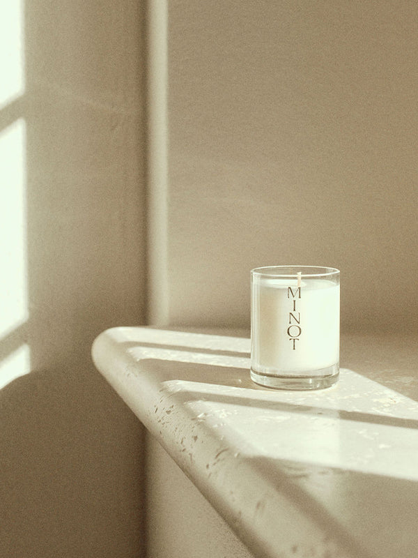 A MINOT mini candle in a clear glass holder is placed on a sunlit ledge, casting soft shadows on a textured wall.