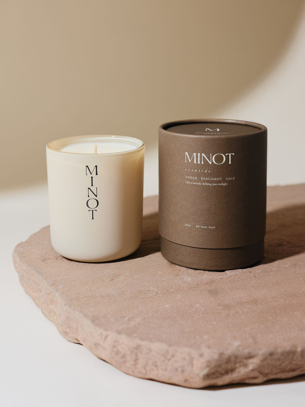 A non-toxic soy candle in a glass vessel labeled "MINOT" sits on a textured stone surface next to a brown cylindrical box labeled "Eventide: Amber, Bergamot, Sage"