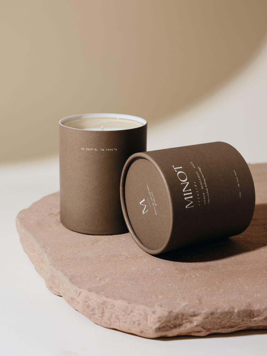 Brown cylindrical packaging holds a MINOT Eventide scented candle with its lid tilted to the side on a textured stone slab. The background is a soft beige color.