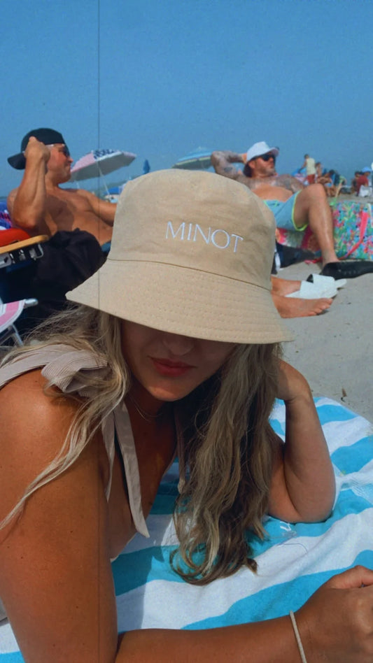 A cream-colored bucket hat with "MINOT" in white embroidery shields a blonde's eyes at the beach