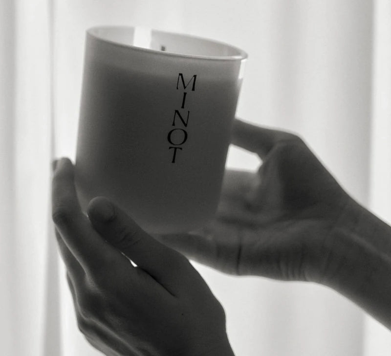 Two hands hold a minimalist, soy wax MINOT candle up toward a sunlit curtain in black and white