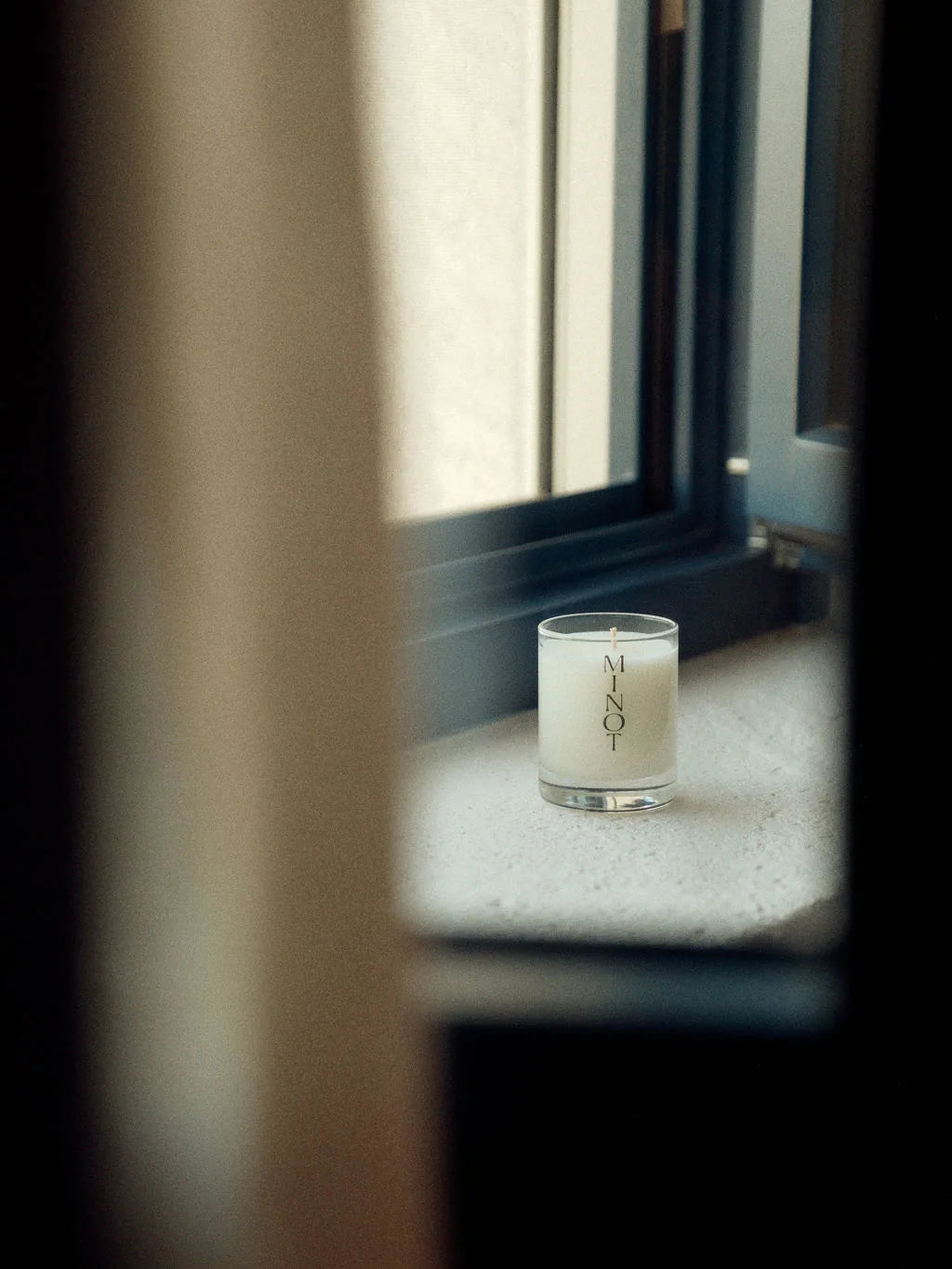 MINOT's Cliffwalk mini candle sits on a window ledge, emanating a sandalwood-patchouli scent blend