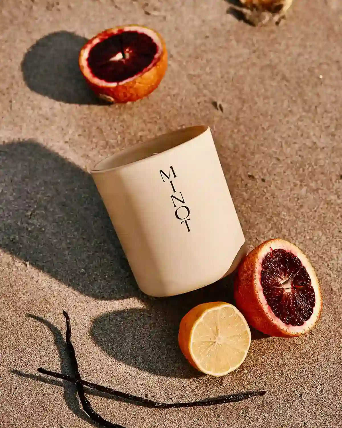 The Sunbeam freesia candle is nestled in beach sand next to vanilla beans, lemons, and blood oranges