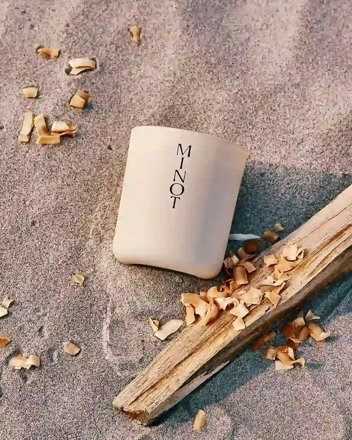 The natural Voyage candle lays in beach sand and coconut shavings next to a piece of driftwood