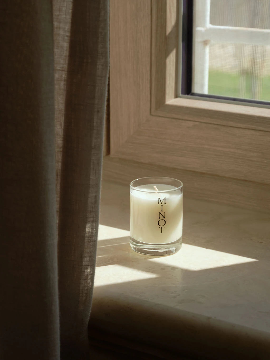 Made with non-toxic ingredients, the Voyage Mini candle is in a window next to a green linen curtain