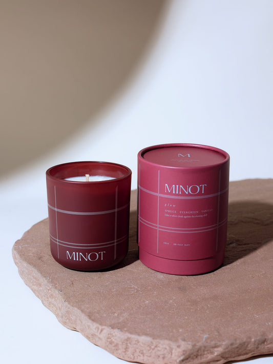 A MINOT scented candle in a dark red glass jar next to its matching cylindrical tin packaging, both featuring a subtle plaid pattern, displayed on a rustic stone slab against a warm beige background.